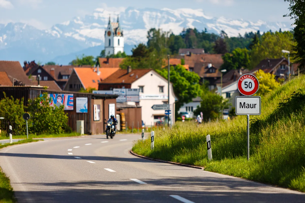 Blick auf eine Strasse mit einer Ortstafel und der Kirche von Maur.